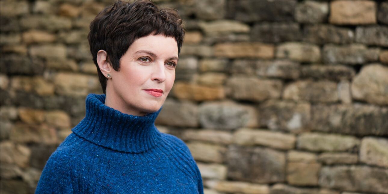 A colour portrait of Louise Tickle in front of a stone wall. Tickle is a white woman with short dark hair wearing a blue polo necked jumper. She faces at angle to the camera.